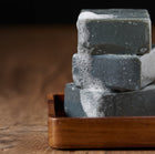 Stack of charcoal soap bars with suds on a wooden tray against a dark background