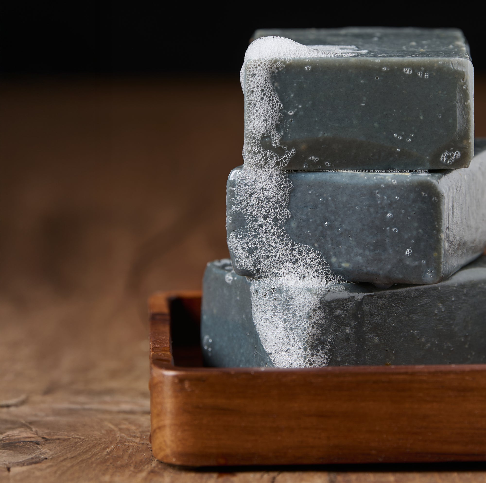 Stack of charcoal soap bars with suds on a wooden tray against a dark background