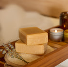 Two bars of soap on a wooden tray with a candle and lavender flowers in the background.