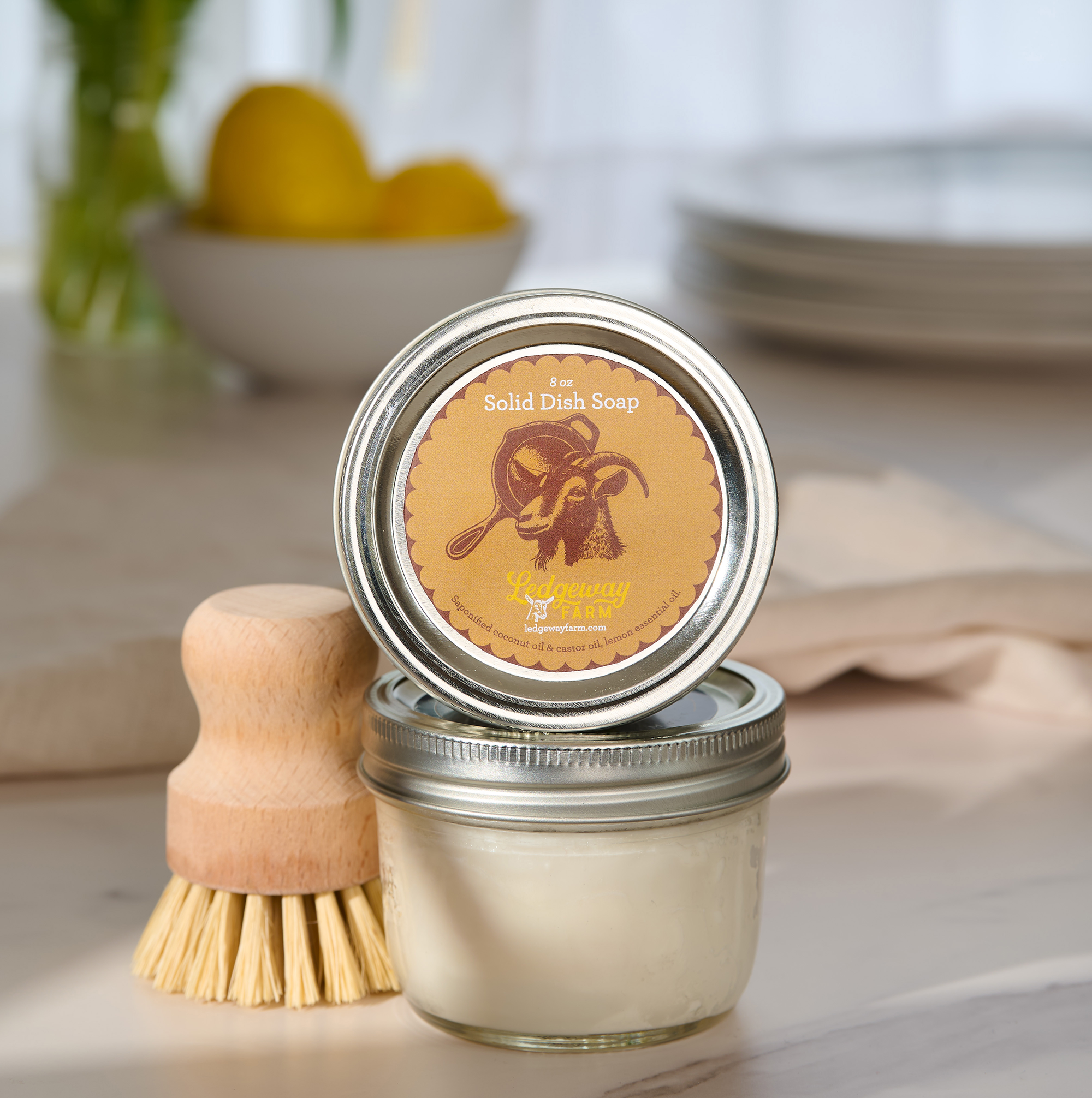 Two jars of solid dish soap with a wooden brush on a kitchen counter.