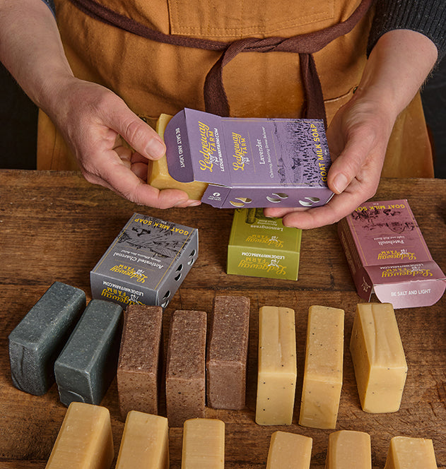 Person holding soap packaging with various soap bars on a wooden surface