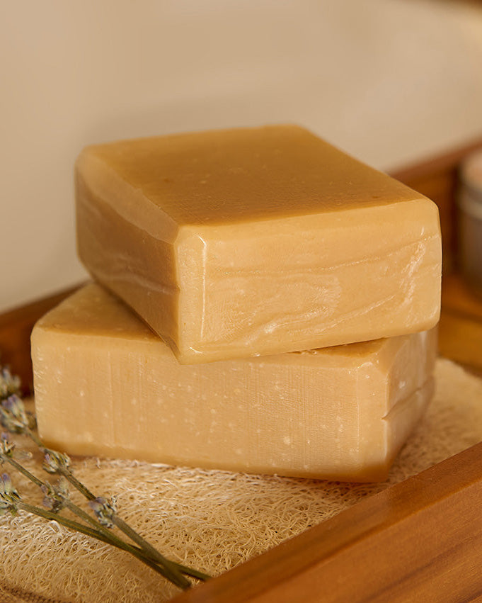 Two bars of soap stacked on a wooden tray with a candle and lavender flowers in the background.