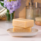 Two bars of soap stacked on a white dish with a blurred background featuring a vase of flowers and bottles.
