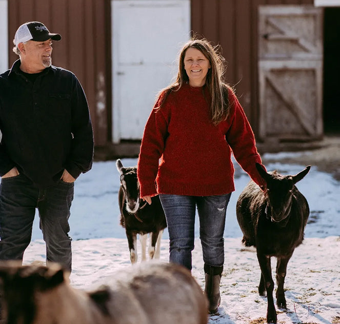 Two people with two goats in a snowy outdoor setting near a barn.