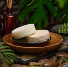 Two white soap bars on a wooden tray with a natural background of rocks and plants.